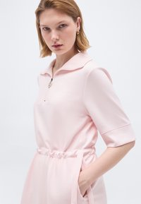 Νεαρή γυναίκα με κοντά ξανθά μαλλιά φοράει ένα ανοιχτό ροζ φόρεμα με φερμουάρ, μανίκια μήκους μέχρι τον αγκώνα, σφιγμένη μέση και χρυσούς κρίκους σκουλαρίκια.