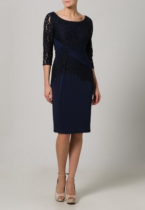 Femme portant une robe bleu marine jusqu'aux genoux avec des manches en dentelle et une superposition de dentelle florale, associée à des chaussures ouvertes à talons hauts beige.