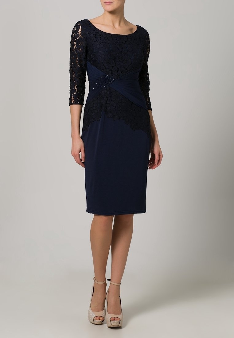 Femme portant une robe bleu marine jusqu'aux genoux avec des manches en dentelle et une superposition de dentelle florale, associée à des chaussures ouvertes à talons hauts beige.