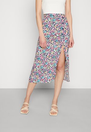 Kobieta w midi spódnicy w kwiaty w różowe, niebieskie i żółte kwiaty, z wysokim rozcięciem na udzie, w połączeniu z białymi sandałami na ramiączkach.