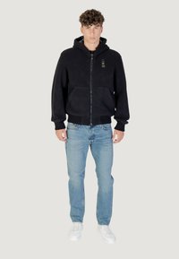 Felpa nera con zip e cappuccio, tasche frontali e polsini a costine, abbinata a jeans azzurri chiari e sneakers bianche.
