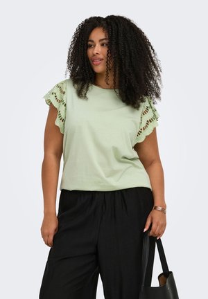 Mujer con cabello rizado que lleva una camiseta verde claro con mangas de encaje y pantalones negros, sosteniendo un bolso negro.