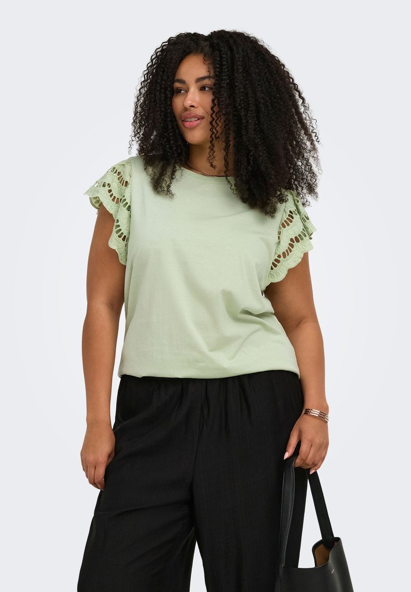 Femme aux cheveux bouclés portant un haut vert clair avec des manches en dentelle et un pantalon noir, tenant un sac à main noir.