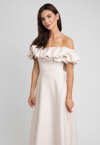 Suknia z odkrytymi ramionami w jasnym beżu, z falbaną przy dekolcie i dopasowanym gorsetem, który przechodzi w swobodnie opadającą spódnicę. Gładka faktura tkaniny.