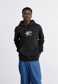 Sort hættejakke med snore og frontlomme, med et hvidt logo på brystet. Kombineret med løstsiddende blå denimjeans.