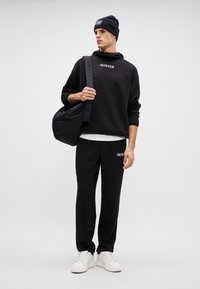 Čierna bavlnená mikina s logom "Calvin Klein", čierne tepláky a biele tenisky. Modelka tiež nesie čiernu kabelku a má na sebe čiernu beanie.