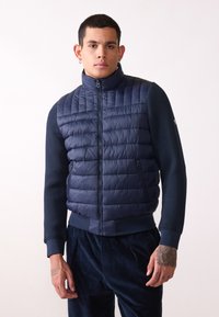 Mörkblå uppblåsbar väst med kviltad textur, hög krage, dragkedjestängning och två sidofickor, bärs över en mörkblå långärmad tröja.