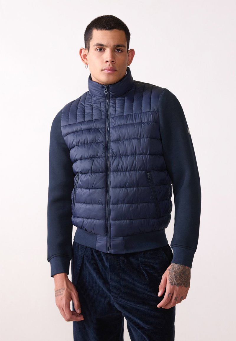 Mörkblå uppblåsbar väst med kviltad textur, hög krage, dragkedjestängning och två sidofickor, bärs över en mörkblå långärmad tröja.