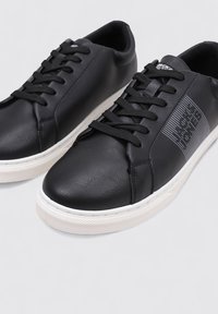 Zapatillas de cuero negro con textura suave, suela de goma blanca y cordones negros. Presentan un sutil detalle gráfico en el lateral.