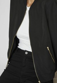 Zwarte bomberjack met gouden ritsdetails, geribbelde manchetten en zoom. Draagt over een wit shirt, gecombineerd met zwarte broek.