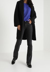 Personne portant un long manteau noir, un pull bleu, un jean noir et des mocassins noirs brillants, debout devant un fond clair uni.