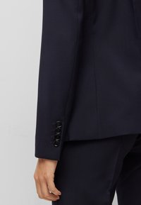 Manica di un blazer navy con quattro bottoni neri, texture liscia, vestibilità sartoriale e dettagli di cucitura sottili, abbinato a pantaloni coordinati.