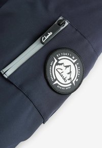 Tejido de color navy con una textura suave que presenta un bolsillo con cremallera. Un parche redondo negro muestra montañas, texto y coordenadas, realzando el diseño.