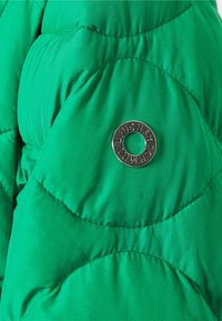 Veste en doudoune verte avec sections matelassées, texture lisse et un logo en métal rond accentuant la zone de la poitrine.