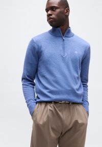 Άνδρας που φοράει μπλε πουλόβερ με φερμουάρ μέχρι τη μέση και το λογότυπο "ck" και μπεζ πλισέ παντελόνι, με τα χέρια στις τσέπες, κοιτάζοντας πλάγια σε απλό φόντο.