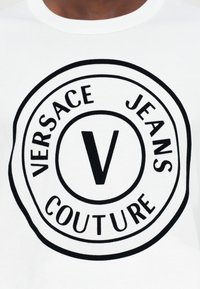 Balts kokvilnas T-krekls ar apaļo melno logo, kurā ir teksts "VERSACE JEANS COUTURE" un centrālais stilizētais "V" dizains.