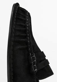 Black suede shoe featuring a textured upper, decorative ruffles, and a flat sole. Closure includes a strap with a subtle woven design.