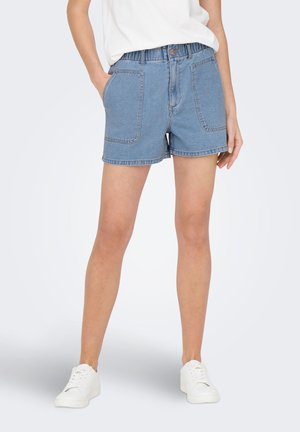 Person iført lyseblå cowboyshorts med elastisk linning og store forlommer, hvide sneakers og en hvid top.