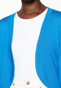Blå cardigan med mjuk, texturerad stickning och öppen frontdesign, bärs över en vit åtsittande t-shirt, kombinerad med vita högmidjade byxor.