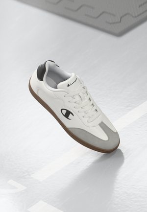 Zapatillas blancas con acentos grises y suelas de goma marrón, con un logo en el costado y paneles texturizados. Diseño con cordones, material de piel suave.