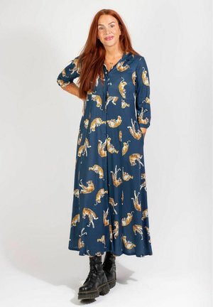 Langes Kleid aus marineblauem Stoff mit einem durchgehenden Tigerprint. Vorn mit Knöpfen, langen Ärmeln und seitlichen Taschen. Getragen mit schwarzen Schnürstiefeln.