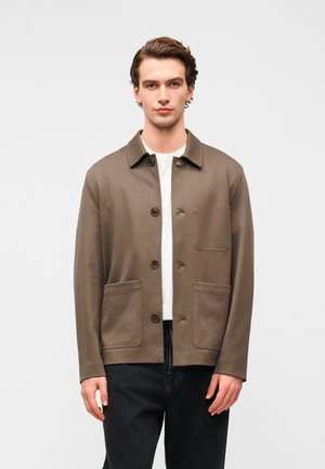 Veste en coton marron avec un col, une fermeture à boutons et deux poches avant. Portée sur une chemise blanche, associée à un jean foncé. Texture lisse.