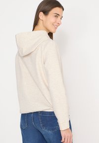 Femme souriante, portant un sweat à capuche beige clair et un jean bleu, légèrement tournée sur le côté devant un fond blanc uni.