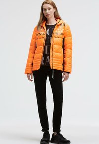 Padded orange jacket with zip closure and printed accents on one side, paired with a black graphic t-shirt, black pants, and black sneakers.