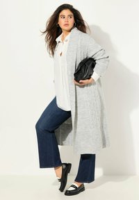 Šedý pletený kardigan s ribkovanou strukturou, bílá blůza, tmavé džínové zvonové kalhoty a černé slip-on boty s bílými akcenty.