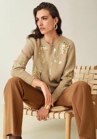 Frau sitzt auf einem geflochtenen Lederstuhl, trägt einen beigefarbenen Pullover mit goldenen, blumigen Pailletten, braune weite Hosen, goldene Creolen und ein Armreif.