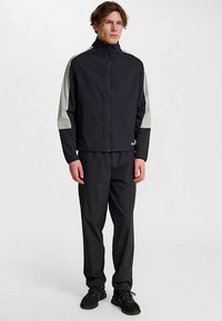 Schwarze Jacke mit Reißverschluss und grauen Ärmelakzenten, kombiniert mit schwarzen Hosen und dunklem Schuhwerk. Der Stoff wirkt glatt und leicht.