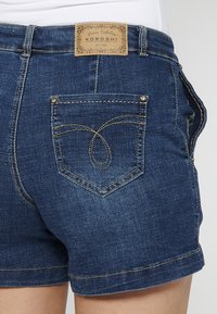 Denim kratke hlače v temno modri barvi z gladko teksturo. Imajo svetlo rjavo usnjeno oznako, zapleteno šivanje zadnjega žepa in kovinske gumbne dodatke.