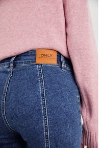 Blue denim jeans feature a brown leather label with "ONLY" text at the waistband, paired with a light pink knit sweater.