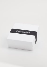 Balts taisnstūra kaste ar melnu joslu, kurā ir teksts "Calvin Klein" baltā fontā. Gluda tekstūra, minimālistisks dizains.