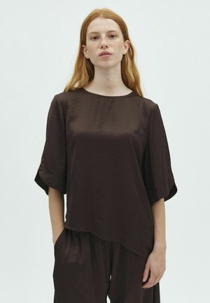 Jeune femme aux longs cheveux roux portant un chemisier ample marron foncé à manches courtes et un pantalon assorti, sur un fond clair uni.