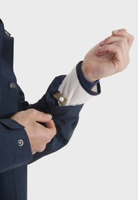 Giacca impermeabile blu con dettagli ai polsini a bottone a pressione e fodera in pile visibile al polsino; presenta una vestibilità strutturata e una texture liscia.
