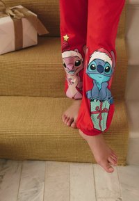 Pantaloni del pigiama rossi con personaggi dei cartoni animati e accenti festivi. Una gamba presenta un personaggio con un cappello di Babbo Natale, mentre l'altra ha un design a scatola regalo.