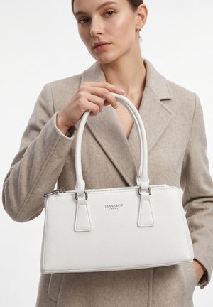 Woman in a beige coat holding a white Diana&Co Firenze handbag with dual handles against a plain white background.