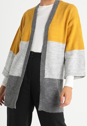 Kobieta ubrana w kardigan w kolorach blokowych: musztardowy żółty, jasnoszary i ciemnoszary, na białej prążkowanej koszulce i czarnych spodniach.