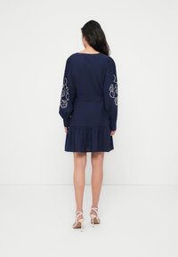 Φόρεμα σε ναυτικό μπλε χρώμα με μακριά μανίκια και κεντημένα λουλουδάτα σχέδια, εφαρμοστή μέση και φουσκωτή φούστα. Το ύφασμα φαίνεται λείο με ελαφριά υφή.