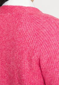 Rosa stickad cardigan med en texturerad yta, avslappnad passform och ribbade detaljer längs kragen och sömmarna.