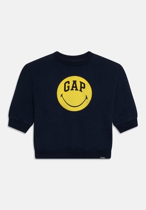 Sweat-shirt bleu marine avec un grand visage souriant jaune et le mot « GAP » en lettres noires et grasses sur la poitrine.
