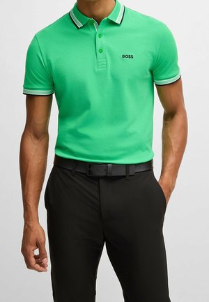 Homme portant un polo manches courtes vert vif avec le logo "BOSS" et un pantalon noir avec une ceinture noire, les mains détendues le long du corps.