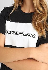 Černé bavlněné tričko s bílým blokem, na němž je tučným černým písmem napsáno "CALVIN KLEIN JEANS". Krátké rukávy a kulatý výstřih.