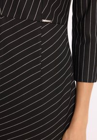 Czarna sukienka w białe paski, dopasowany krój, rękawy do łokcia oraz subtelny metaliczny akcent z logo po boku.
