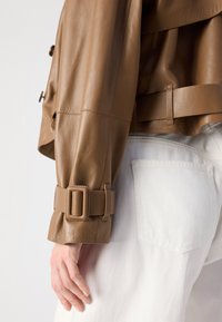 Een close-up van een bruine leren jacksleeve met een gesp en een hand die tegen witte denim leunt, waardoor stijlvolle details en textuur worden benadrukt.