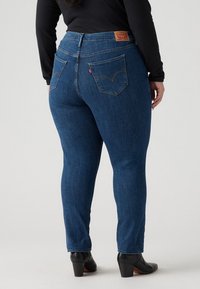 Jeans de mezclilla azul con un ajuste de altura media, que cuentan con bolsillos traseros, un parche de logo de cuero y una textura suave. Combinados con zapatos de tacón negros.