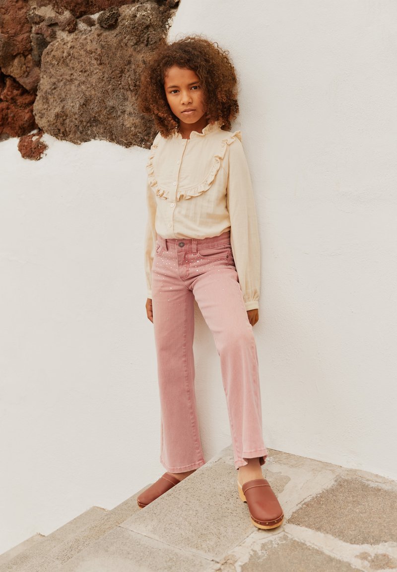 Dieťa s kučeravými vlasmi, oblečené v krémovej blúzke s volánmi, ružových džínoch s ozdobami a hnedých drevákoch, stojí proti bielej kamennej stene.