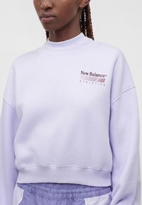 Γυναίκα φοράει ανοιχτό μωβ φούτερ New Balance Athletics με ψηλό γιακά και αντίστοιχο μωβ αθλητικό παντελόνι.