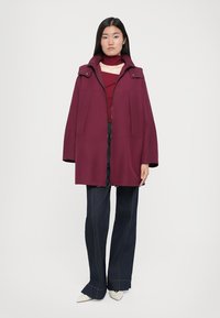 Une femme se tient debout, portant un manteau bordeaux sur un haut assorti, avec un jean large noir et des chaussures blanches à bout pointu, devant un fond clair.
